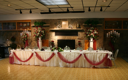 Wedding Table in Front of Fireplace with Red, Green and White Floral centerpiece as well as flowers framing fireplace, red tulle draped from table