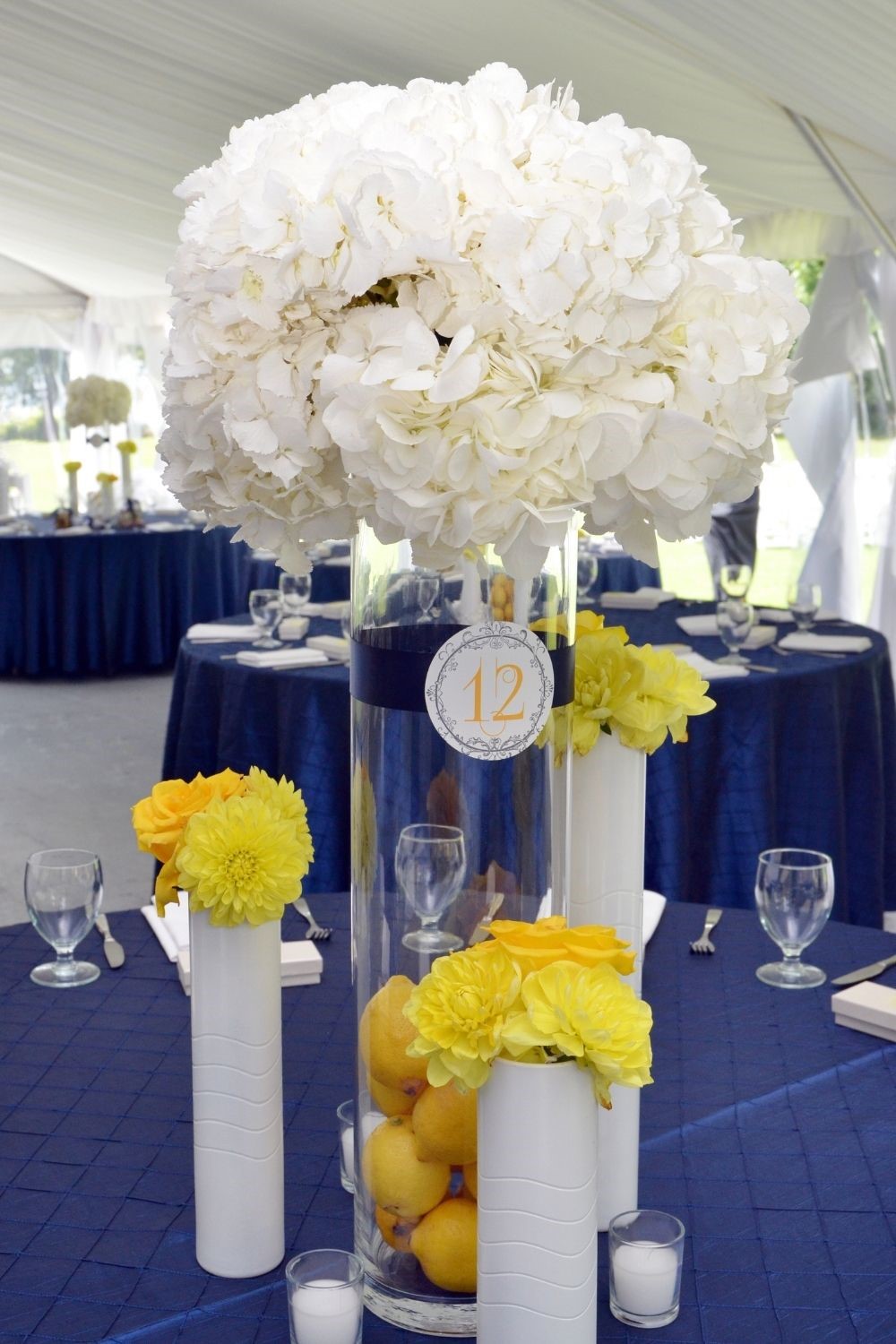 Large White Floral Wedding Centerpiece in vase with lemons surrounded by white candles and smaller yellow floral arrangements in white vases